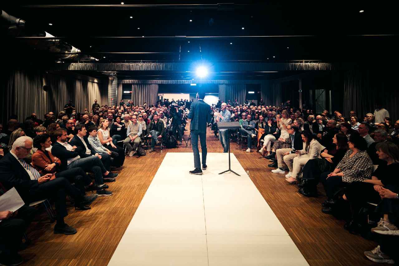 Matteo Biffoni parla, sul palco, davanti a una sala gremita di sostenitori. La foto, scattata dal fondo della sala, lo mostra di spalle, circondato da persone sedute su entrambi i lati. 
